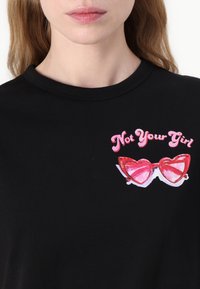 Musta T-paita, jossa on vaaleanpunaista tekstiä "Not Your Girl" punaisen sydänmuotoisen aurinkolasikuvaston päällä, jossa on valkoisia yksityiskohtia ja heijastuksia.