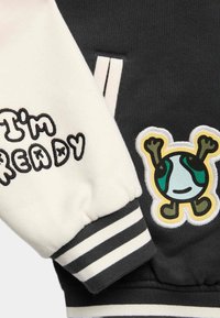 Mikina s černo-krémovým barevným schématem, s pruhovaným manžetovým lemem a výšivkami, včetně veselého charakteru a textu.