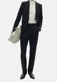 Costume noir sur mesure avec pantalon slim, un col roulé gris clair et des chaussures noires pointues. Un sac à main gris clair complète le look.