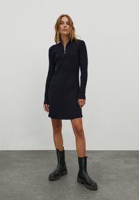 Marineblaues geripptes Strickkleid mit hohem Kragen und Reißverschlussdetail, ausgestattet mit langen Ärmeln und einer geraden Silhouette, kombiniert mit schwarzen klobigen Stiefeln.