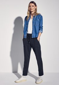 Camisa de mezclilla azul con cierre de botones, camiseta blanca de tirantes y pantalones jogger negros con bolsillos laterales. El calzado son zapatillas blancas.