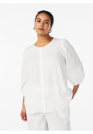 Kobieta w luźnej białej bluzce z trzy-czwarte rękawami typu bufki oraz dopasowanych białych spodniach, stojąca z jedną dłonią w kieszeni.