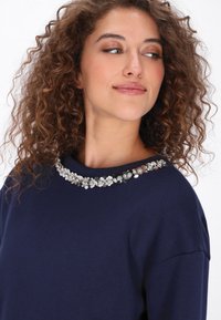 Bluza de culoare albastru marin, cu decolteu decorat cu mărgele florale. Material moale din bumbac, cu croială lejeră, mâneci lungi și design cu guler rotund.