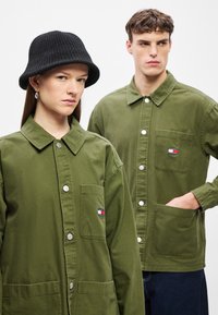 Χακί βαμβακερά σακάκια με κουμπιά μπροστά και τσέπες, λευκά κουμπιά και μικρά μπαλώματα με λογότυπο. Μία μοντέλο φοράει μαύρο ριμπ καπέλο τύπου bucket.