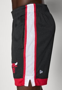 Shorts en mesh noirs avec des bandes latérales rouges et blanches, dotés d'un logo brodé et d'un ourlet fendu en bas, conçus pour le confort et la respirabilité.