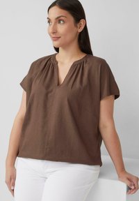 Bruine korte mouwen blouse met een V-hals, plooidetail bij de halslijn en een losse pasvorm, gecombineerd met witte broek tegen een neutrale achtergrond.