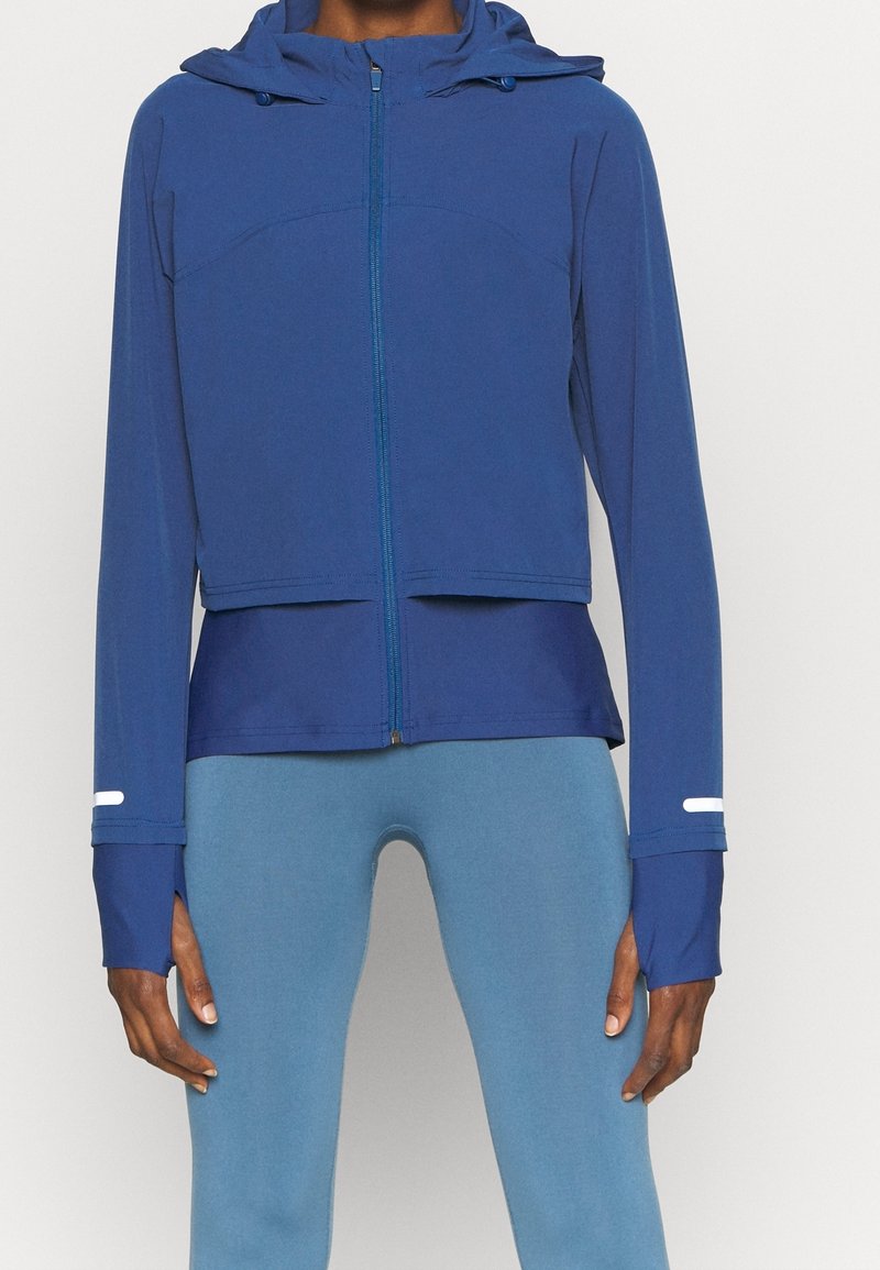 Blå sportjacka med dragkedja fram, huva och långa ärmar. Har tumhål och en cropped design. Tillverkad av ett slätt, stretchigt material.