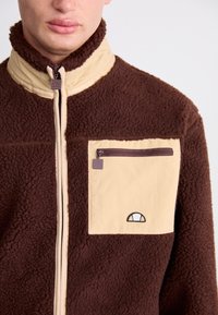 Veste en polaire marron avec des accents beiges, col montant, fermeture éclair à l'avant et une poche sur la poitrine. Présente un logo au-dessus de la poche. Texture douce.