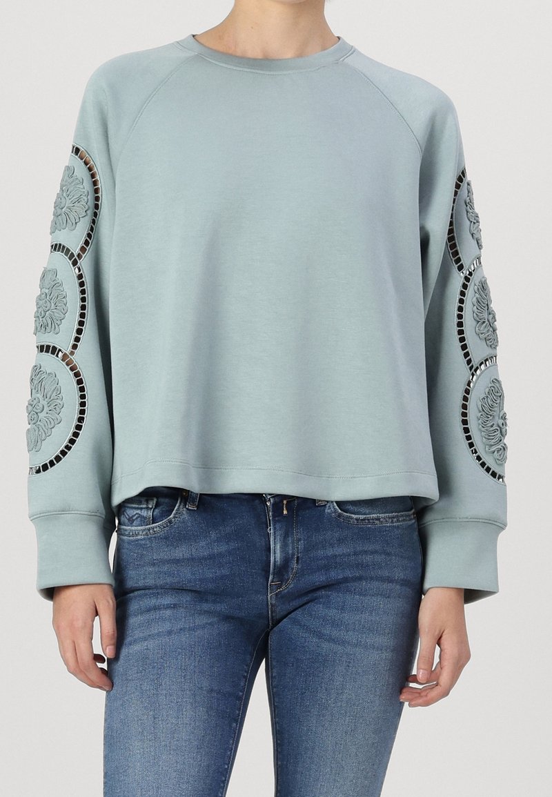 Sweat-shirt bleu clair à manches raglan, avec des motifs floraux brodés sur les manches et un ourlet court. Texture de tissu douce.