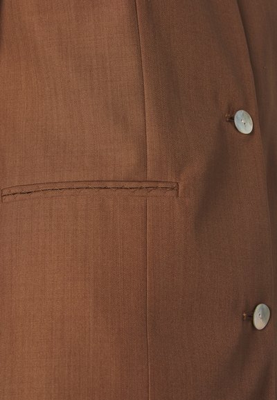 Καφέ σακάκι προσαρμοσμένο με λεία υφή, διαθέτει μια μεμονωμένη τσέπη και ένα μεγάλο κουμπί για κλείσιμο. Ραφές γύρω από την τσέπη.