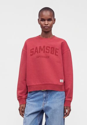Voksen iført en rød sweatshirt med teksten "SAMSØE COPENHAGEN" og lyseblå jeans, stående mod en ensartet lys baggrund.
