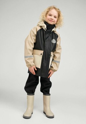 Wasserdichter schwarzer und beiger Regenmantel mit Kapuze, Fronttaschen und reflektierenden Details, kombiniert mit schwarzer Hose und beigen Gummistiefeln.