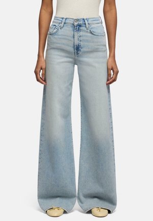 Kvinde iført lyseblå, højtaljede, vidde jeans og beige sko, stående med hænderne afslappet langs siderne mod en hvid baggrund.