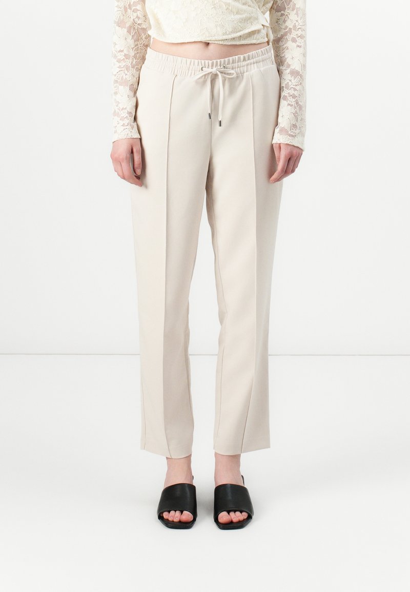 Beige, elastiske bukser med lige ben og foran plissering, parret med sorte slidssandaler. Materialet ser glat og let ud.
