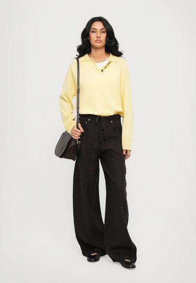 Camisa polo de manga larga amarilla con tres botones, combinada con pantalones negros de pierna ancha. Llevando un pequeño bolso cruzado gris y zapatos negros.