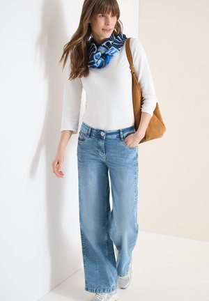 Femme portant un haut blanc à manches longues, un jean bleu à jambes larges, une écharpe bleue à motifs, des baskets blanches et portant un sac à bandoulière marron, debout à l'intérieur.