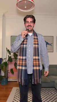 Stripete skjorte med knapper og lange ermer, kombinert med et rutete skjerf i marineblått, brunt og oransje. Sorte bukser, innendørs omgivelser med planter.