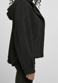 Personne portant une veste à capuche noire texturée avec des manches longues, vue de profil des épaules aux hanches.