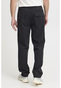 Black cotton trousers with a relaxed fit, featuring a back pocket with a button closure and tapered legs. Worn with light-colored sneakers.