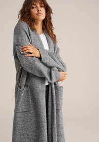 Garšs atvērts kardigans mīkstā pelēkā adījumā, ar lielām kabatām un ribotām piedurknēm. Audumam ir teksturēta virsma.