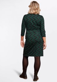 Vihreä kuvioitu mekko, jossa on vartalonmyötäinen siluetti, kolmanneksen mittaiset hihat ja teksturoitu kangas, yhdistettynä mustiin sukkahousuihin ja nilkkureihin.