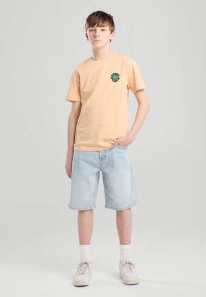 Camiseta de algodón durazno con cuello redondo que presenta un diseño de globo verde bordado; combinada con pantalones cortos de mezclilla azul claro y zapatillas blancas.