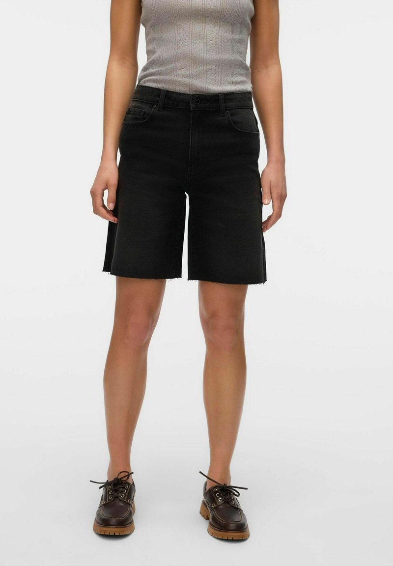 Pantalones cortos de denim negro con dobladillo sin terminar, con una cintura alta y bolsillos frontales. Llevados con una camiseta sin mangas gris y zapatos marrones de cordones.