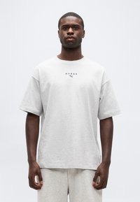 Νεαρός μαύρος άνδρας που φοράει ανοιχτό γκρι T-shirt HYROX Puma και αντίστοιχα σορτς, στέκεται μπροστά σε απλό λευκό φόντο.