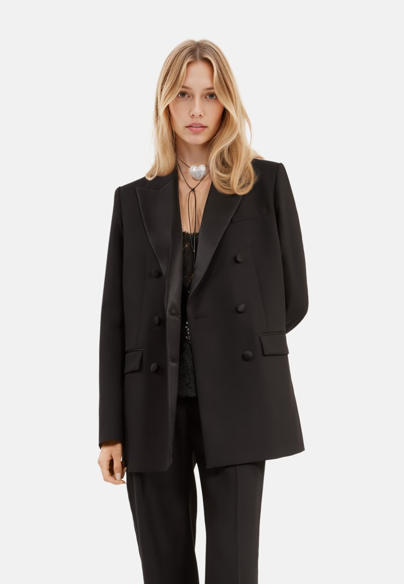 Blazer doppiopetto nero con finitura satinata, revers a punta e grandi bottoni. Indossato su una blusa in pizzo nero trasparente e abbinato a pantaloni coordinati.