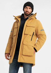 Geel geïsoleerde parka met meerdere zakken, een ritssluiting aan de voorkant en een capuchon. Heeft grijze reflecterende accenten op de mouwen. Draagt over een marineblauwe hoodie.