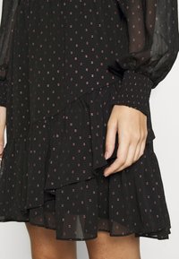 Vestido negro con estampado que presenta un diseño en capas, tejido transparente con pequeños puntos rosas y puños elásticos. El dobladillo con volantes añade textura.