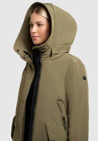 Olivgrüner Parka mit großer Kapuze, Reißverschluss und seitlichen Taschen. Besitzt eine glatte Textur und schwarze Akzente an den Reißverschlüssen und Knöpfen.
