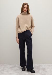 Beige Rollkragenpullover aus weichem Material, kombiniert mit dunkelblauen, ausgestellten Jeans. Das Outfit wird durch schwarze klobige Schuhe ergänzt.