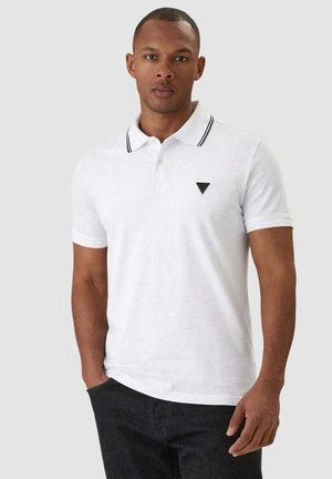 Uomo che indossa una polo bianca con colletto bordato di nero e logo triangolare nero, in piedi con una mano in tasca su sfondo grigio.