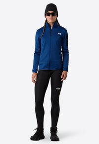 Modrá zipová bunda vyrobená z měkkého materiálu, spárovaná s černými legínami a černými botami. Doplňky zahrnují černou čepici a sluneční brýle.