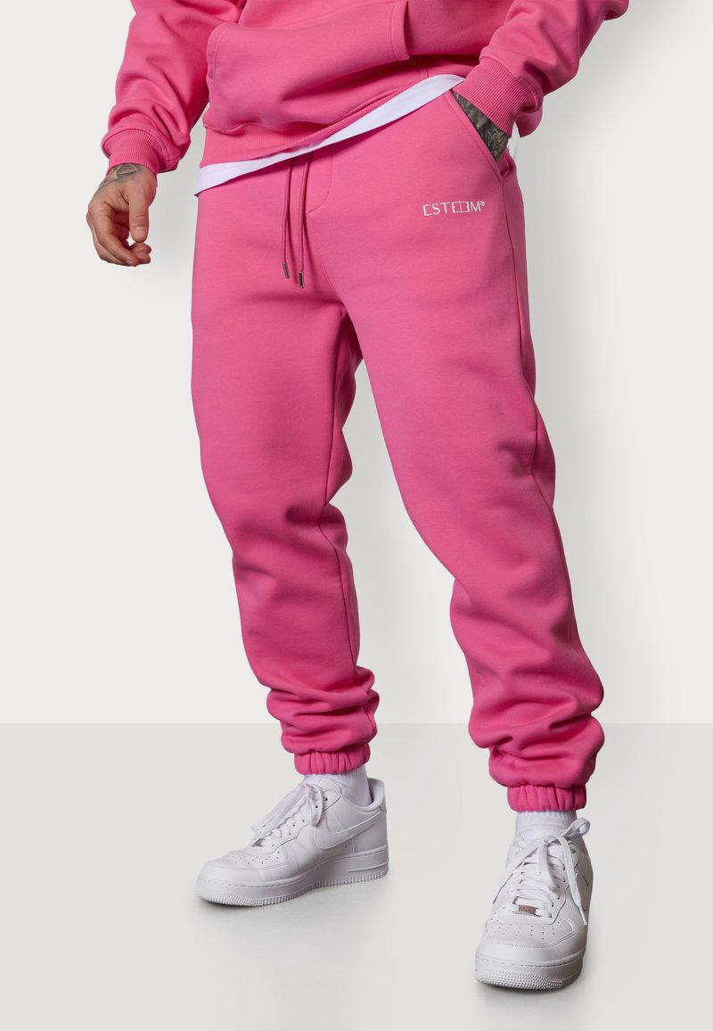 Rosa Jogginghose aus weichem Stoff mit elastischem Bund und Bündchen, ausgestattet mit einer Vorderentasche und einem Logo-Detail am Oberschenkel. Weiße Sneakers.