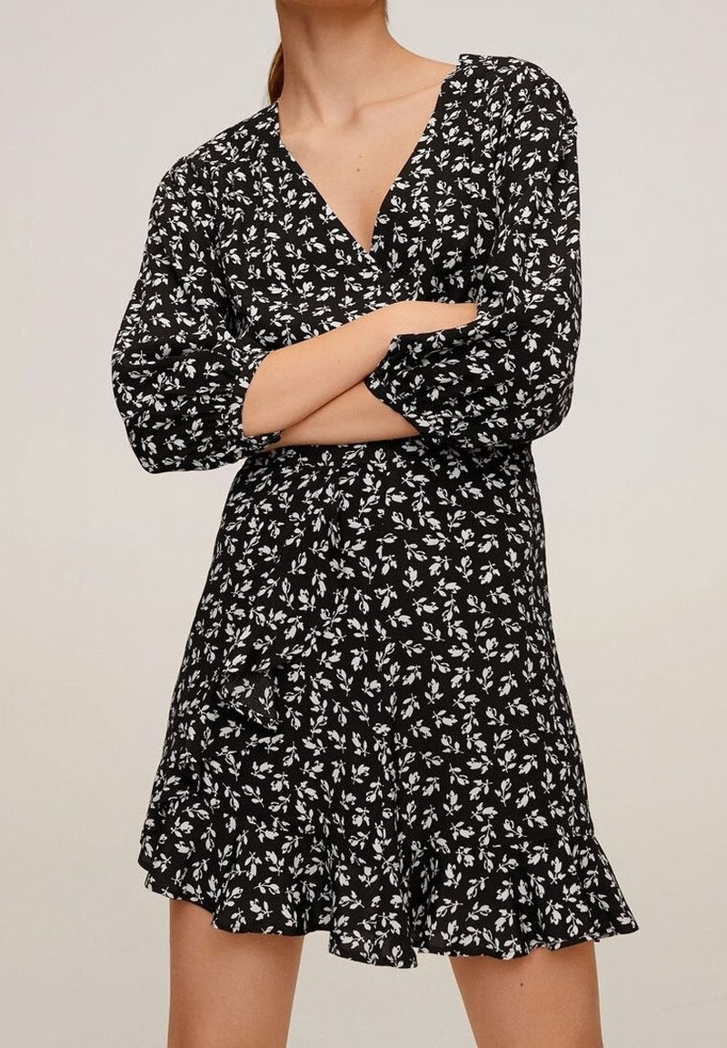Femme portant une robe noire à imprimé floral blanc, col en V, manches longues et ourlet à volants, bras croisés sur la poitrine.