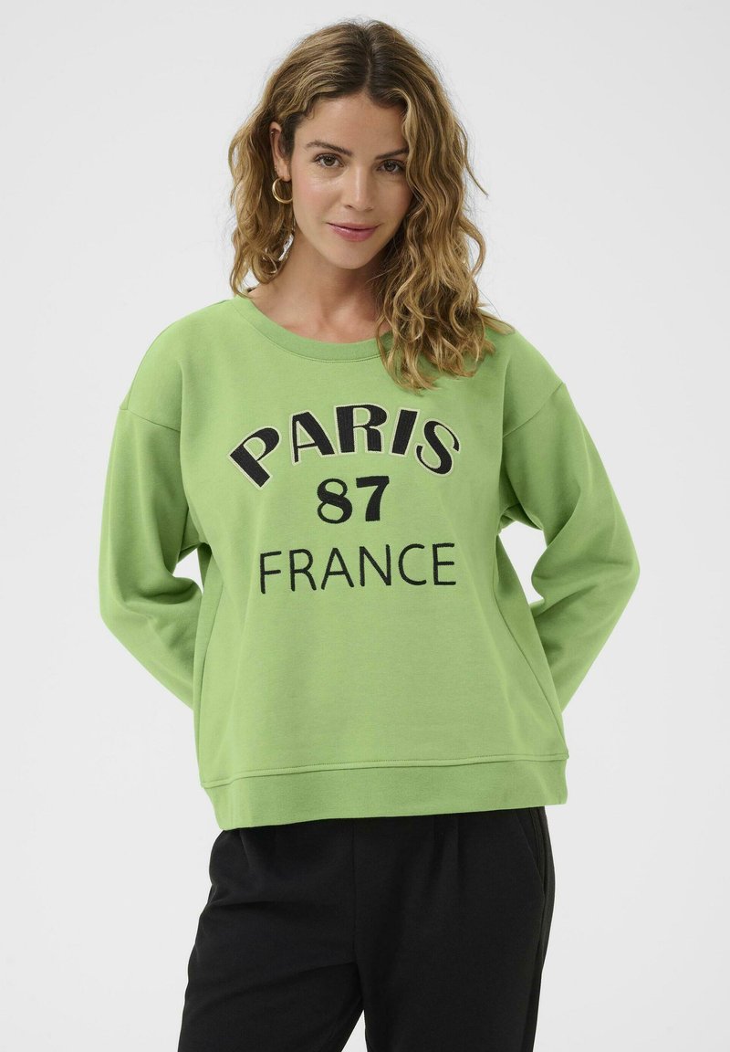 Donna con capelli ondulati, che indossa orecchini a cerchio dorati, una felpa verde con la scritta "PARIS 87 FRANCE" e pantaloni neri, in piedi davanti a uno sfondo bianco.