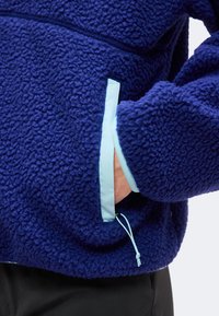 Άτομο που φοράει μπλε ζακέτα fleece με ανοιχτό μπλε τσέπη και κρατάει το χέρι του μέσα στην τσέπη.
