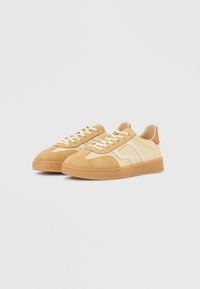 Tan suede sneakers with cream accents, white laces, and a textured rubber sole. Features a simple design and minimal branding on the heel.