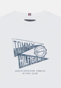 Weißes Baumwoll-T-Shirt mit blauem Grafikdesign und "TOMMY HILFIGER"-Schriftzug sowie Namen von Standorten in NYC. Rundhalsausschnitt und kurze Ärmel.