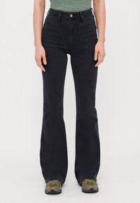Jeans de mezclilla negros de cintura alta y campana, con dos bolsillos frontales y cierre de botón de cobre. Llevados con zapatillas color oliva.