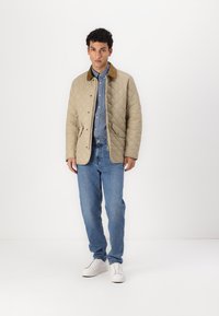 Beige quilted jacket with brown collar, paired with a blue shirt and light blue jeans. White sneakers complete the outfit.