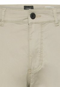 Beige broek vooraan in close-up met zwarte knoop, ceintuurlussen, ritssluiting en Camel merklabel aan de binnenkant van de tailleband.