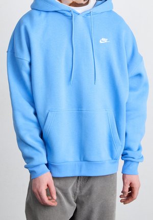 Person trägt einen hellblauen Nike-Hoodie mit Fronttasche und Kordelzug, kombiniert mit grauen Cordhosen, vor einfachem Hintergrund.