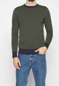 Tumma vihreä neule, jossa on kuvioitu pintakuvio, pyöreä pääntie ja kontrastiväriset tummansiniset hihansuut. Yhdistetty sinisiin farkkuihin.