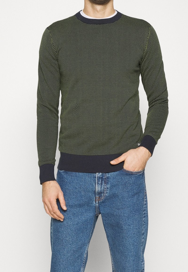 Tumma vihreä neule, jossa on kuvioitu pintakuvio, pyöreä pääntie ja kontrastiväriset tummansiniset hihansuut. Yhdistetty sinisiin farkkuihin.