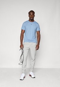 Lysblå atletisk t-shirt med et lille logo, grå sweatpants og hvide sneakers med røde detaljer. Holder en sort sjippetov.