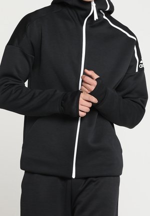 Personne portant un sweat à capuche noir avec une fermeture éclair blanche et un logo Adidas sur l'épaule, les mains ajustant la zone de la fermeture éclair.