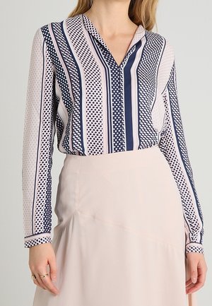 Vrouw die een blouse met lange mouwen draagt met marineblauwe en witte verticale strepen en stippen, gecombineerd met een lichtbeige rok.